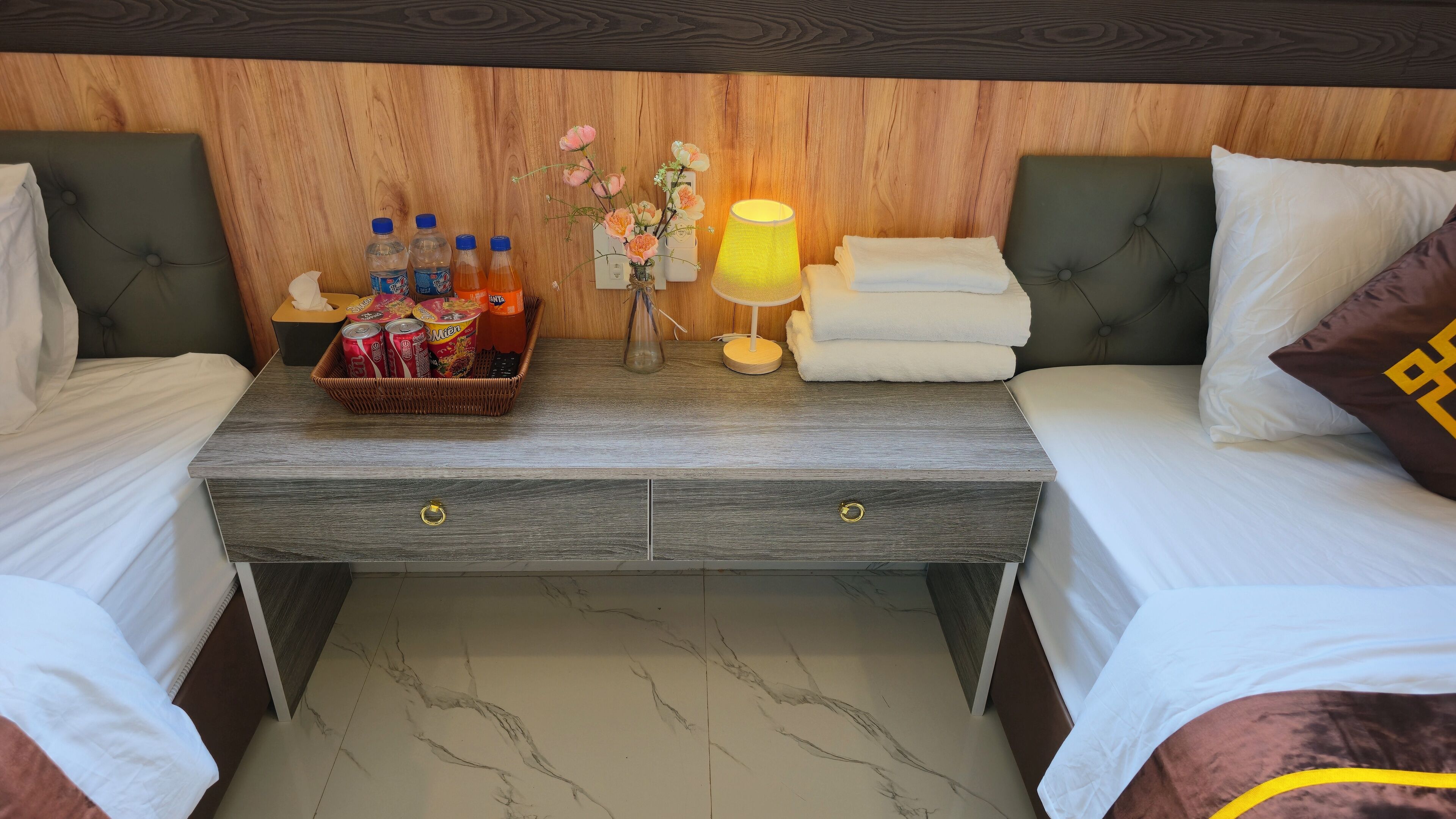 Camera d'albergo con due letti, una testiera in legno, un comodino con una lampada, un cesto di snack e un vaso con fiori.
