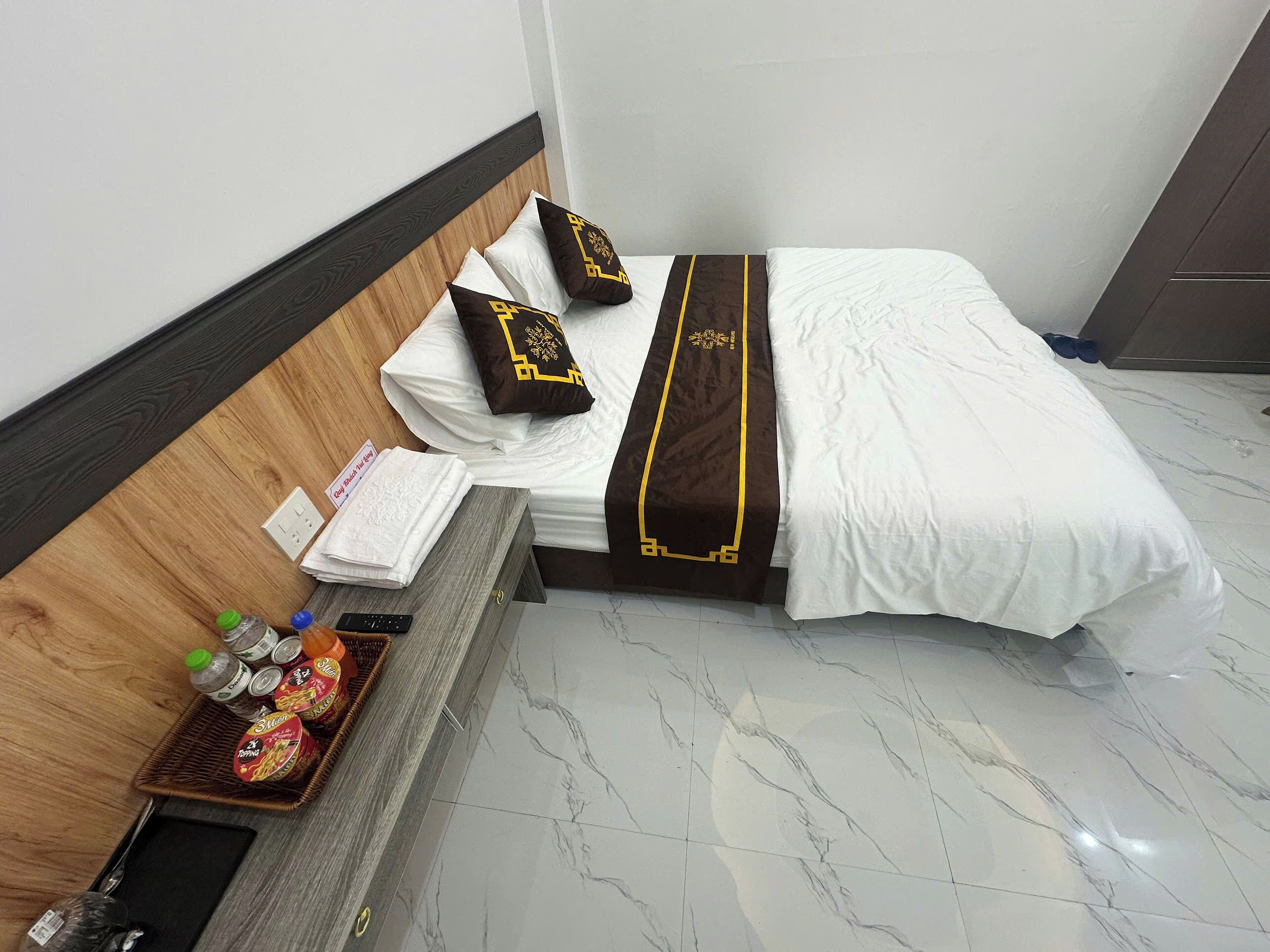 A neatly made bed with white and brown bedding, a headboard, and a bedside table with a basket of snacks and drinks.