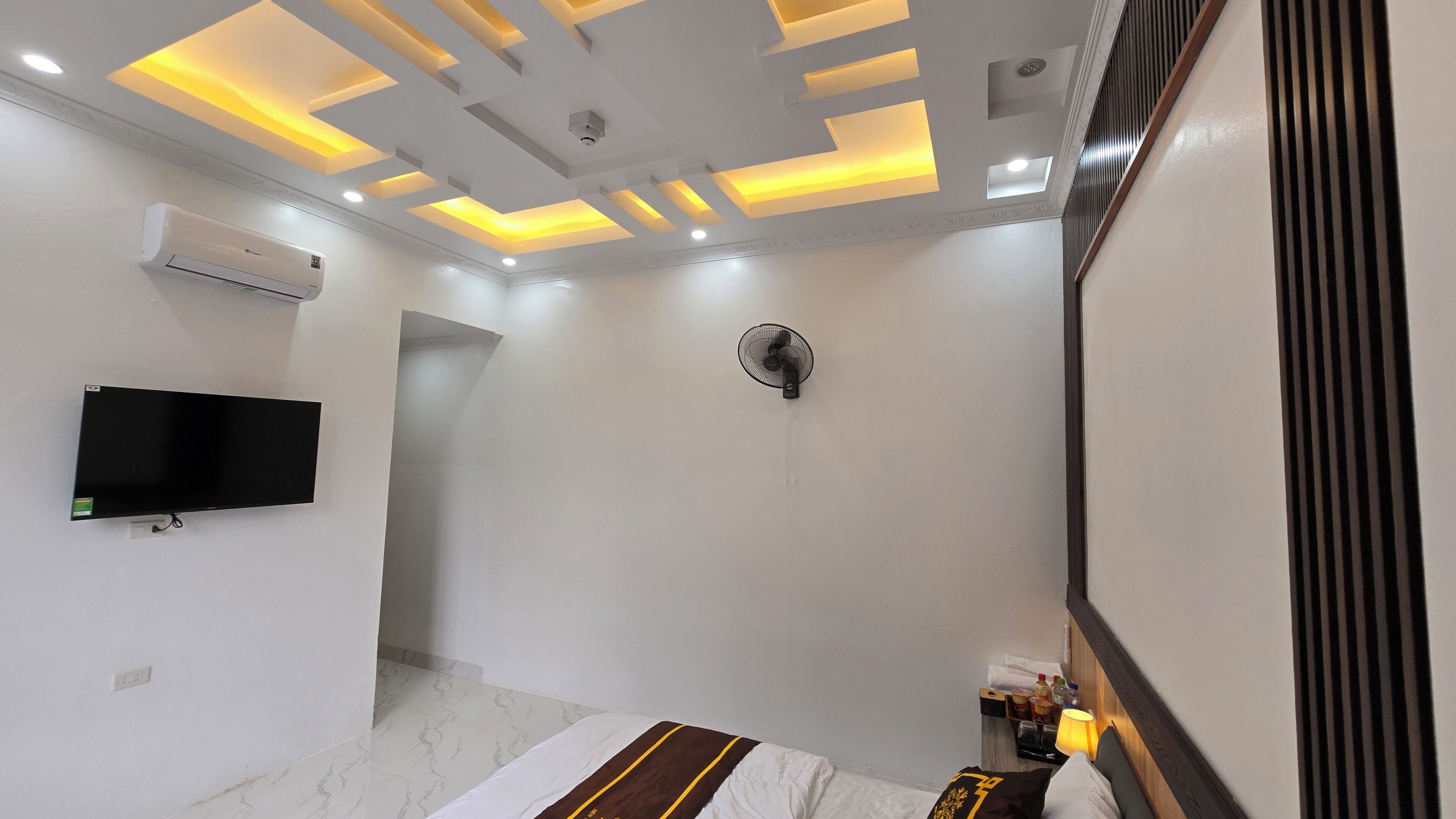 A modern hotel room with a flat-screen TV, air conditioning unit, ceiling fan, and a bed with a striped bedspread.