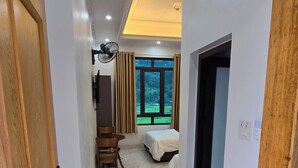 A hotel room with a bed, a desk, a chair, a window with a view of greenery, and a ceiling fan. - New Highland Hotel (Mu Cang Chai)