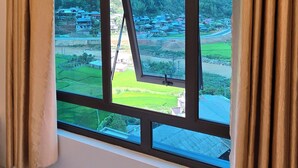 A room with a large window, curtains, and a view of a green landscape with buildings. - New Highland Hotel (Mu Cang Chai)