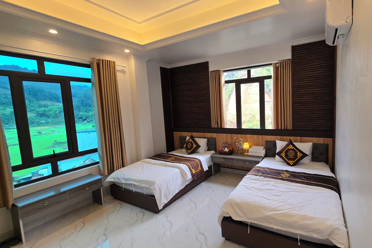 A hotel room with two beds, a window with curtains, a bedside table with a lamp, and a view of greenery outside.