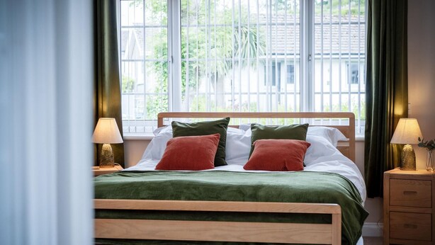 A neatly made bed with a wooden frame, green and red pillows, and a green blanket. There are two bedside tables with lamps and a window with blinds. - Audreys - 3 Bed Holiday Home - Manorbier (Tenby)
