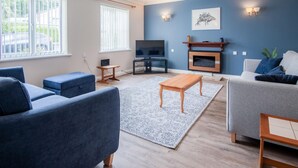 A modern living room with a blue accent wall, a fireplace, and a large window. - Audreys - 3 Bed Holiday Home - Manorbier (Tenby)
