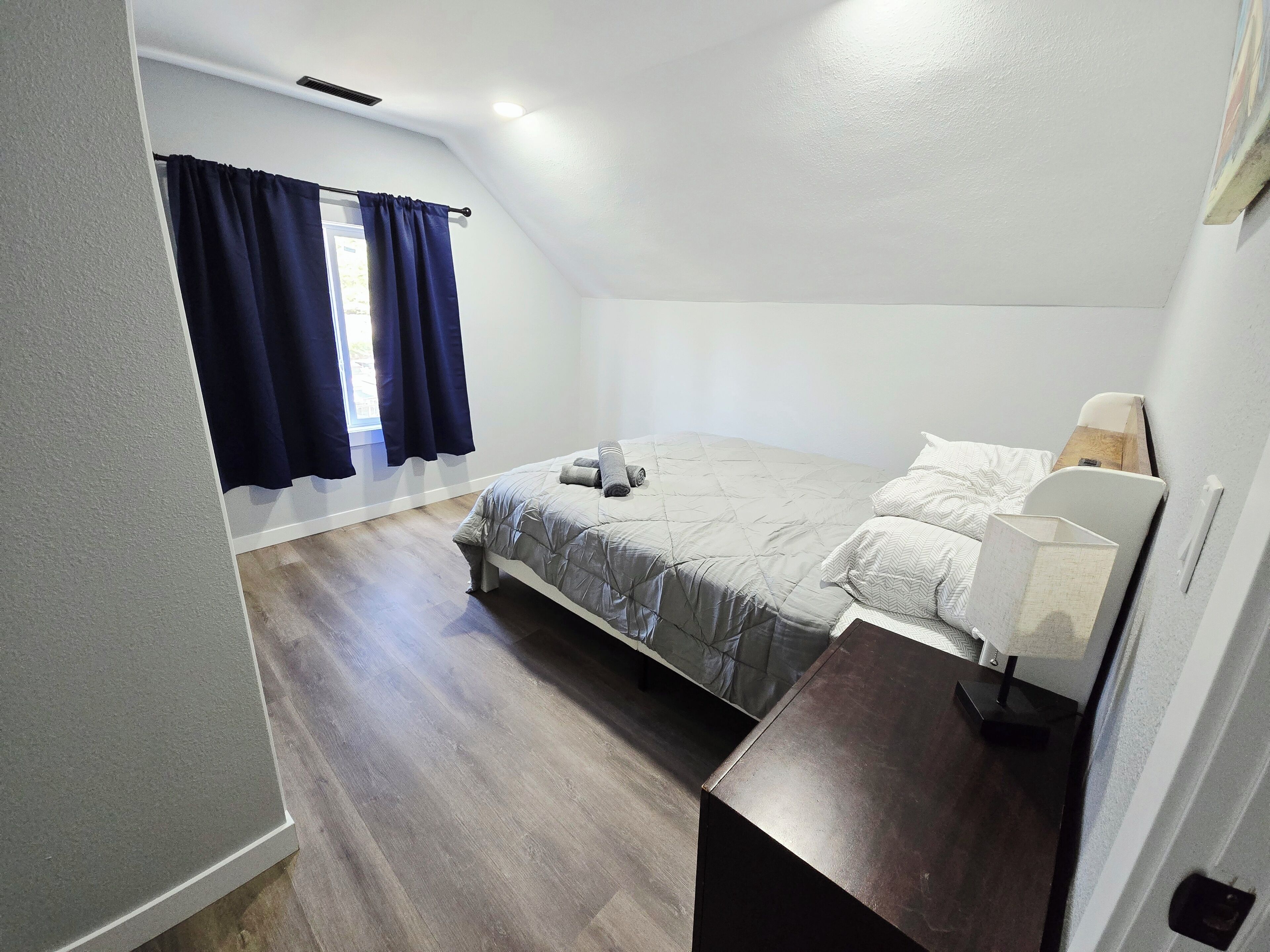 A bedroom with a bed, a bedside table, a window with curtains, and a wooden floor.