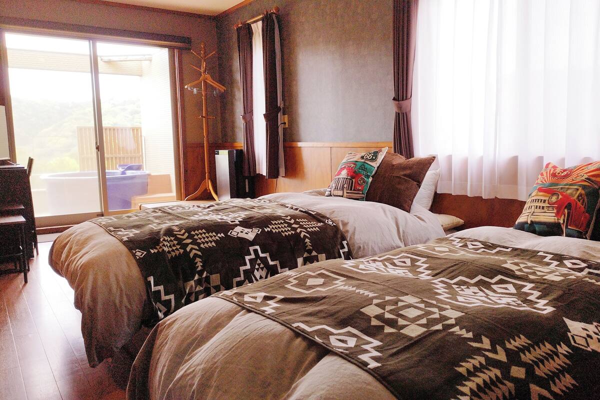A hotel room with two beds, a wooden desk, a chair, a coat rack, and a view of the outdoors through a sliding door.