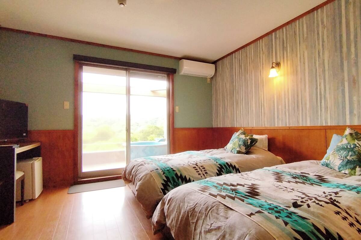 A hotel room with two beds, a TV, a desk, and a sliding glass door to the outside.