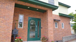 A brick building with a green door, windows, and a small porch area with potted plants. - kokopellihouseizukogen (Ito)
