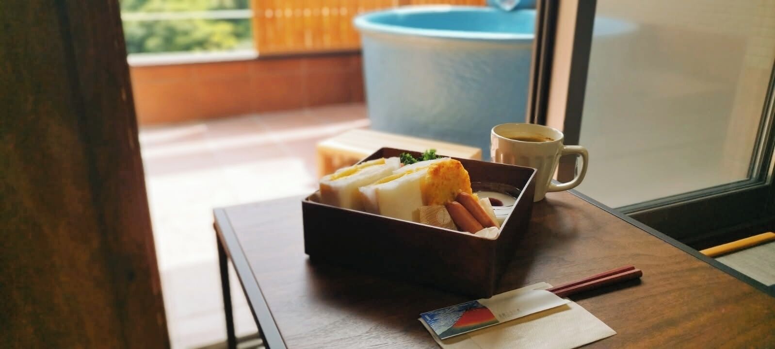 A wooden table with a box containing food, a cup of coffee, and chopsticks.