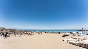 Una extensa playa de arena con sombrillas y sillas de descanso, un promontorio rocoso con un faro, y un cielo azul despejado.