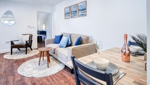 Ein modernes Wohnzimmer mit Sofa, Couchtisch und Esstisch. An der Wand befindet sich ein Spiegel, eine Tür führt in einen anderen Raum und ein kleiner Tisch mit Flasche und Schüssel.