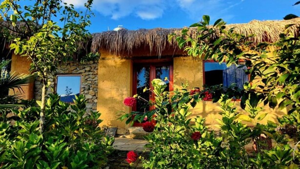 A rustic building with a thatched roof, surrounded by lush greenery and flowering plants. - Homestay Khám Phá Y Tý (Y Ty)