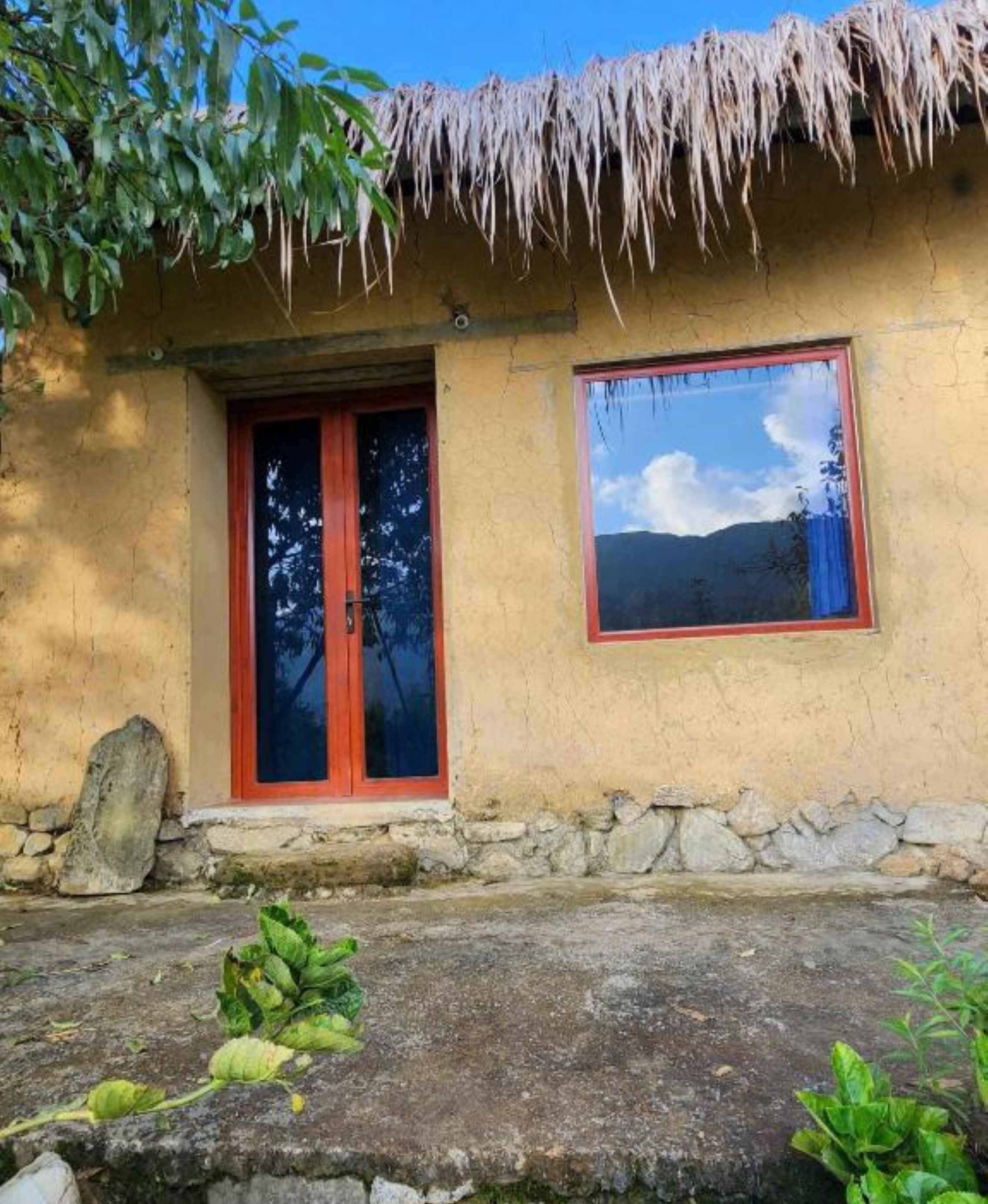 A rustic building with a thatched roof, a red door, and a window reflecting the sky.