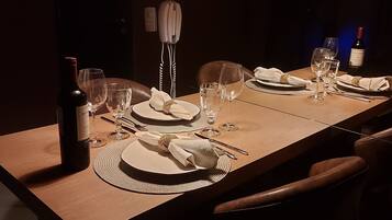 Un comedor con poca luz, con una larga mesa de madera dispuesta para dos personas, que incluye una botella de vino, copas y servilletas dobladas con cuidado.
