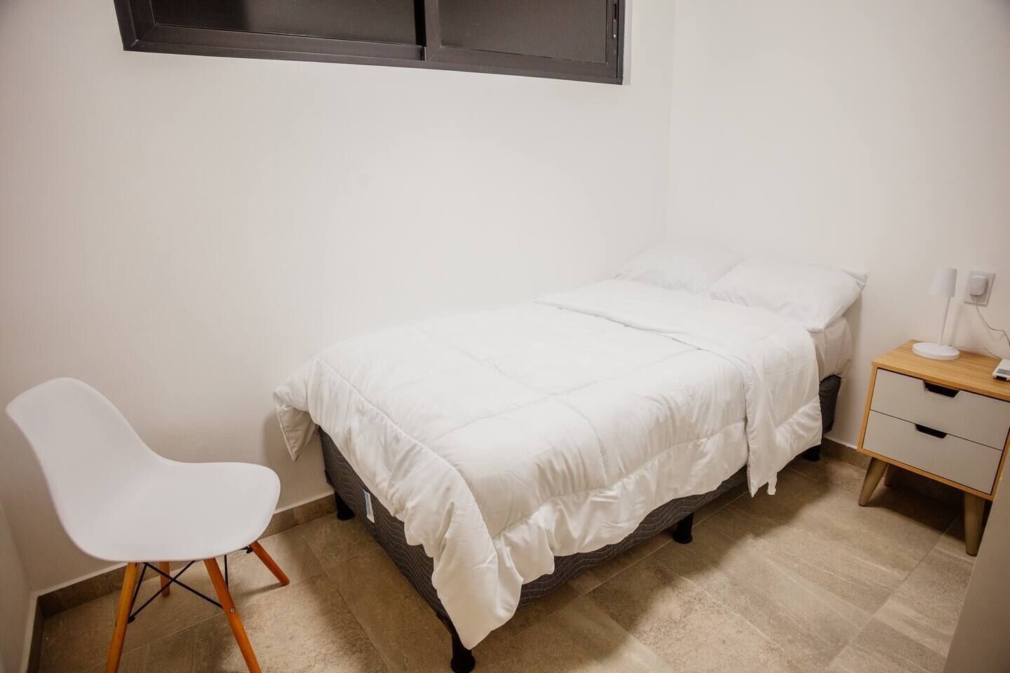 A small, neatly arranged bedroom with a single bed, a bedside table with a lamp, a chair, and a window above the bed.