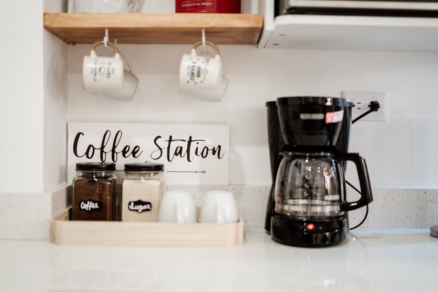 Una estación de café con un cartel, frascos etiquetados como 'café' y 'azúcar', y una cafetera sobre una encimera.