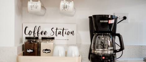 Una estación de café con un cartel, frascos etiquetados como 'café' y 'azúcar', y una cafetera sobre una encimera.