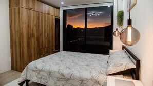 A bedroom with a large wardrobe, a bed with floral bedding, a nightstand, and a view of a sunset through a sliding door.