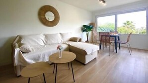 Uma sala de estar moderna com um sofá, duas mesinhas redondas, uma área de jantar com cadeiras, um ventilador de teto e uma grande janela com vista para o verde.