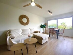 Ein modernes Wohnzimmer mit einer Couch, zwei kleinen runden Tischen, einem Essbereich mit Stühlen, einem Deckenventilator und einem großen Fenster mit Blick auf Grünflächen.