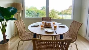 Uma área de refeições com uma mesa redonda, quatro cadeiras de vime, um vaso com planta e vista para o verde lá fora.