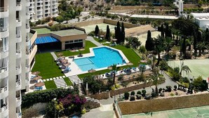 Um resort com uma piscina grande, cercada por espreguiçadeiras e guarda-sóis, com vista para edifícios modernos e uma paisagem acidentada.