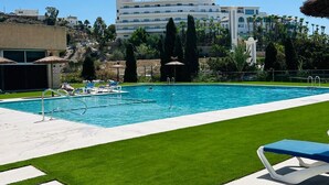 Piscina com água azul cristalina, rodeada por espreguiçadeiras brancas e um gramado bem cuidado.