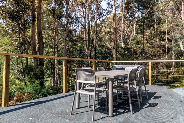 An outdoor dining area with a table and chairs, surrounded by a wooden railing and a forest backdrop.