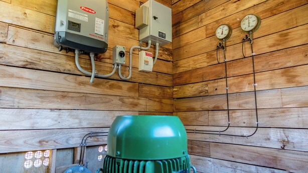 L’intérieur d’une cabane en bois, avec un générateur vert, un appareil de chauffage Fronius et deux manomètres fixés au mur.