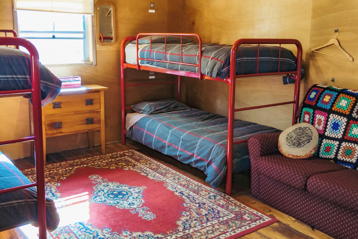 A bunk bed room with a red metal frame, a wooden dresser, a red patterned rug, a purple sofa, and a colorful crochet blanket.