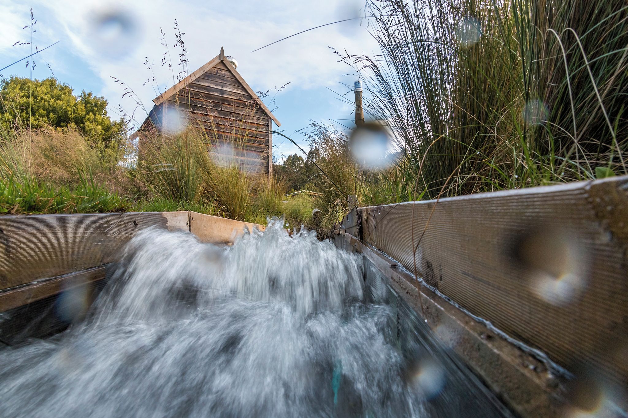 A wooden bridge over a flowing stream with a rustic building in the background.