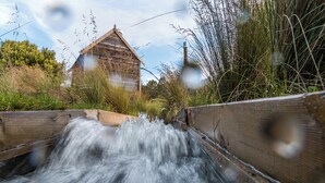 Eine Holzbrücke über einen kleinen Bach, im Hintergrund ein rustikales Gebäude.