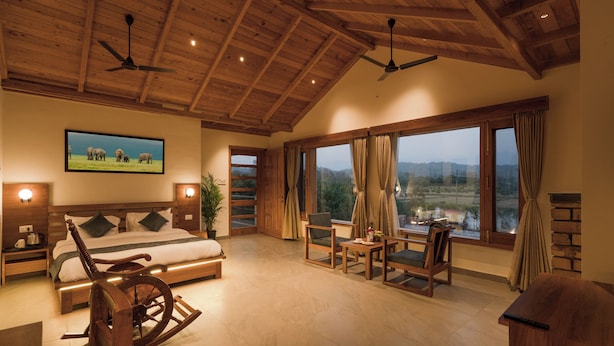 A spacious bedroom with a large bed, wooden furniture, a rocking chair, and a view of a landscape through large windows. - Quinta Premium Resort, Corbett (Kaladhungi)