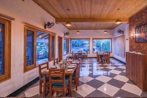 A dining area with checkered flooring, wooden tables and chairs, and large windows offering views of the outdoors. - Quinta Premium Resort, Corbett (Kaladhungi)