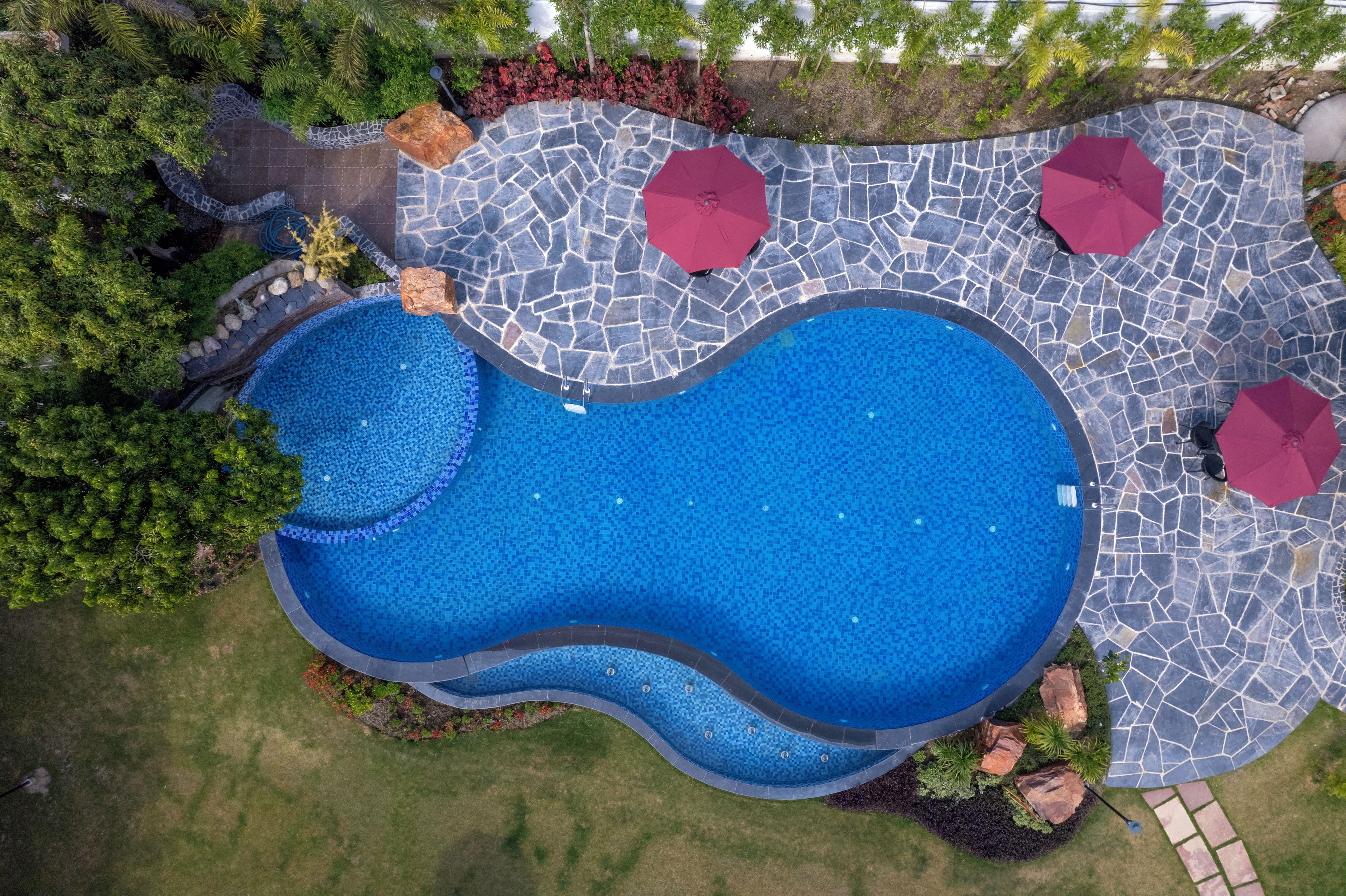 An aerial view of a swimming pool with a unique wavy design, surrounded by a stone patio and red umbrellas.