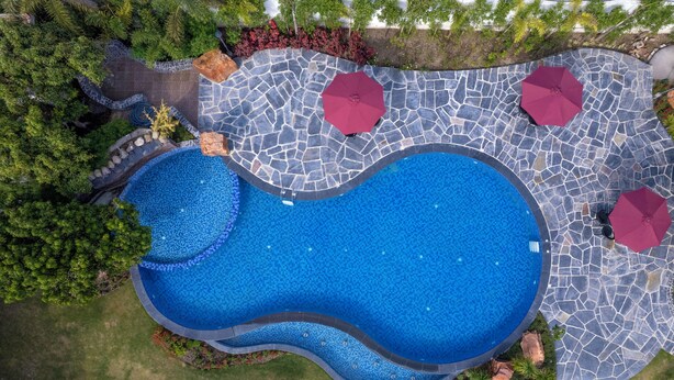 An aerial view of a swimming pool with a unique wavy design, surrounded by a stone patio and red umbrellas. - Quinta Premium Resort, Corbett (Kaladhungi)