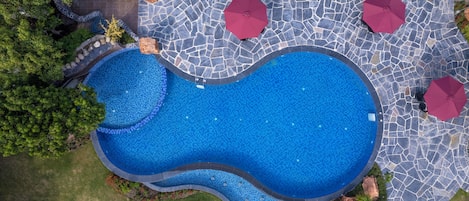 Vista aérea de una piscina con un diseño ondulado singular, rodeada por un patio de piedra y sombrillas rojas.