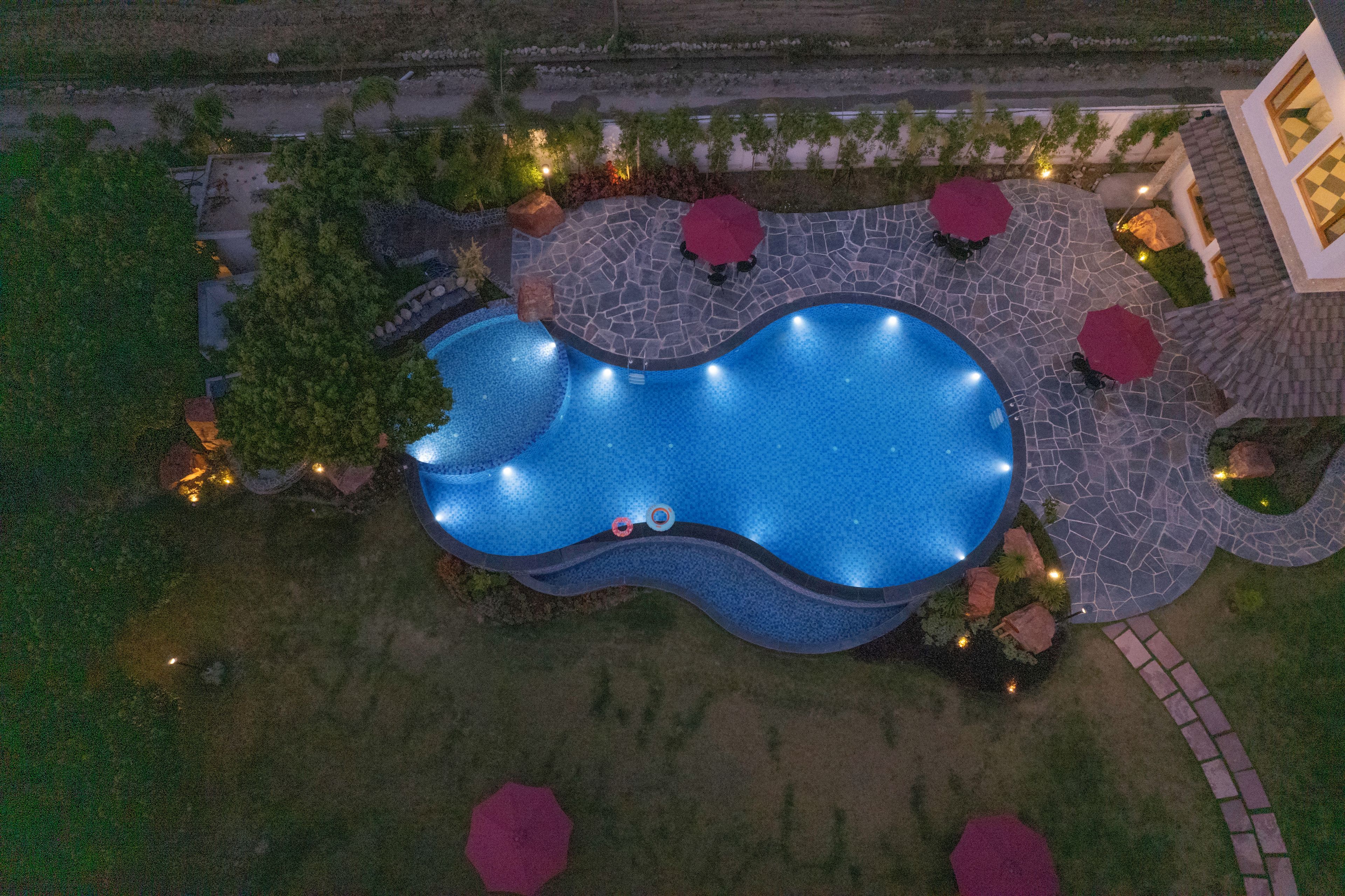 An aerial view of a swimming pool with a stone deck, surrounded by trees and outdoor seating areas.