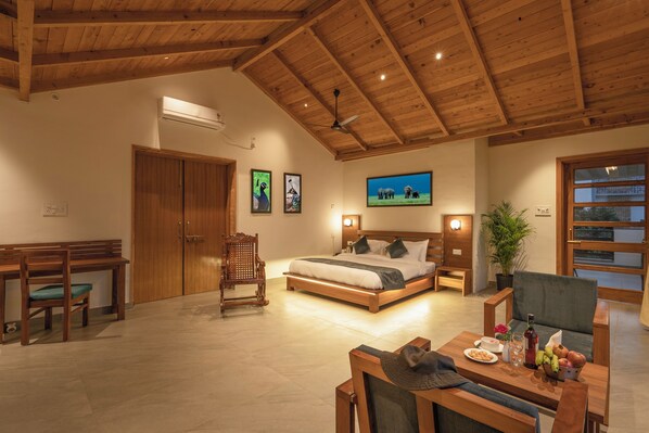 A spacious room with a wooden ceiling, a bed with a headboard, a desk, a chair, and a dining table with fruits and a bottle.