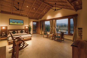 A spacious bedroom with a large bed, wooden furniture, a rocking chair, and a view of a landscape through large windows. - Quinta Premium Resort, Corbett (Kaladhungi)