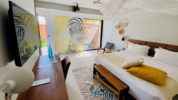 A room with a bed, a desk, a chair, a TV, and a window with a view of greenery. - Hôtel Boma (Dakar)