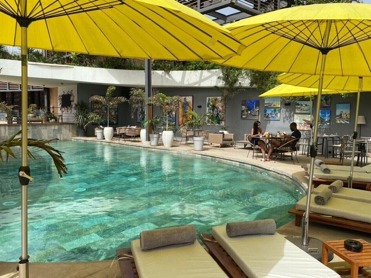 A pool area with lounge chairs, yellow umbrellas, and a restaurant with patrons.
