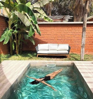A swimming pool with a person swimming, a white sofa, and a brick wall with banana plants. - Hôtel Boma (Dakar)