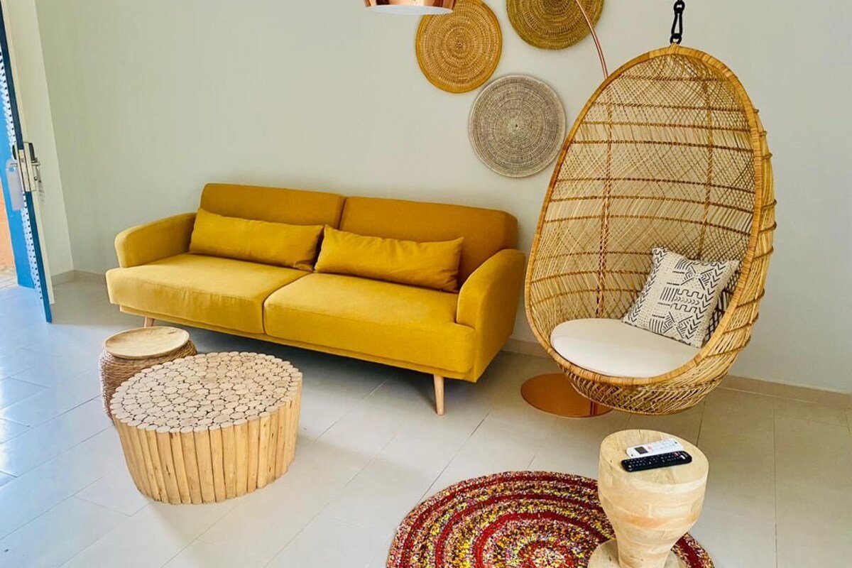 A yellow sofa, a hanging chair, and a round wooden table in a room with a tiled floor.