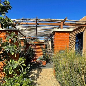 A narrow pathway flanked by brick walls and potted plants, leading to a shaded area with a wooden pergola. - Hôtel Boma (Dakar)