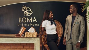A reception area with a sign for BOMA Lifestyle Hotel, a wicker chair, a small table with a tissue box, and a brick wall. - Hôtel Boma (Dakar)