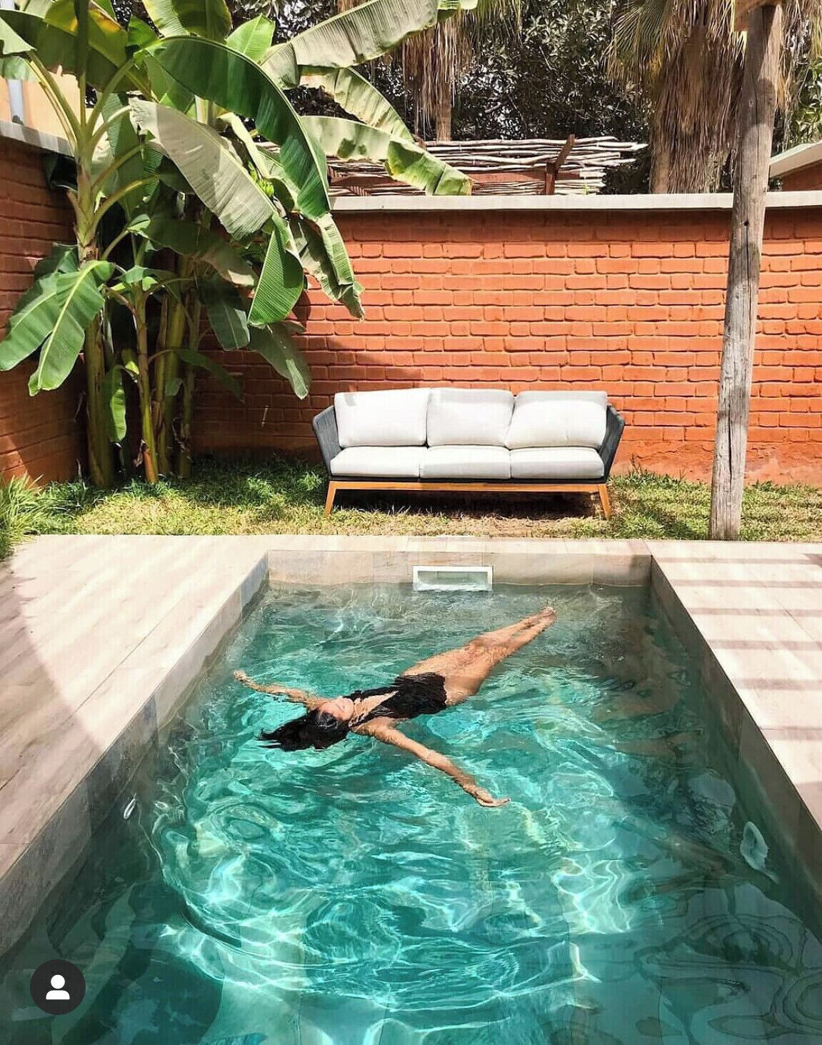 A swimming pool with a person swimming, a brick wall, a white sofa, and banana plants.