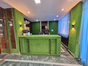 A reception desk with a green artificial grass top, a computer, and a fan. There are two wall-mounted lights on either side of the desk. - Nancy Paradise Hotel (Haridwar)