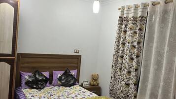 Quarto com cama de madeira, roupa de cama estampada, ventilador de teto e cortinas decorativas.