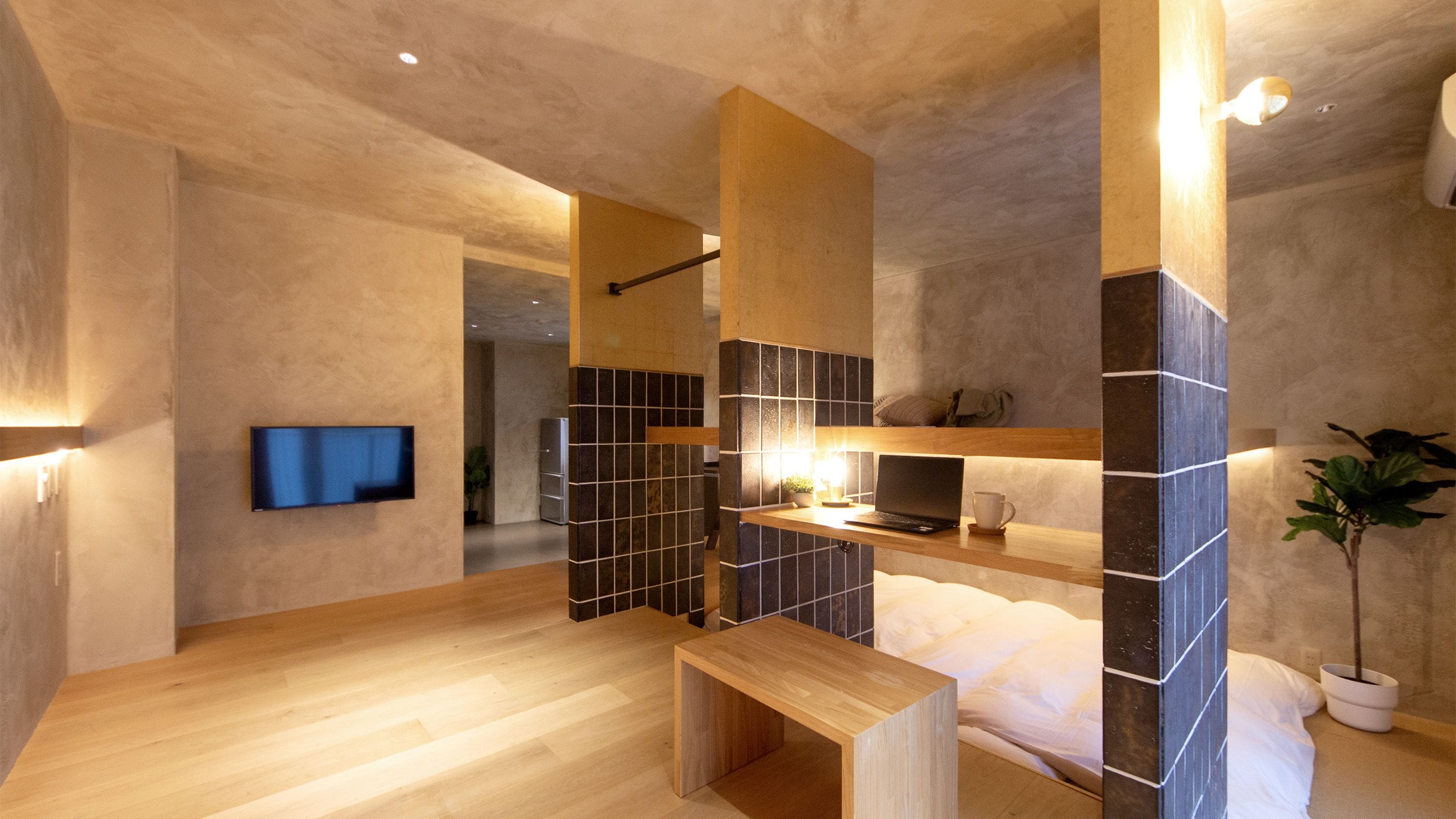 Un baño moderno con un banco de madera, un televisor de pantalla plana montado en la pared y una pequeña mesa con una computadora portátil y una taza.